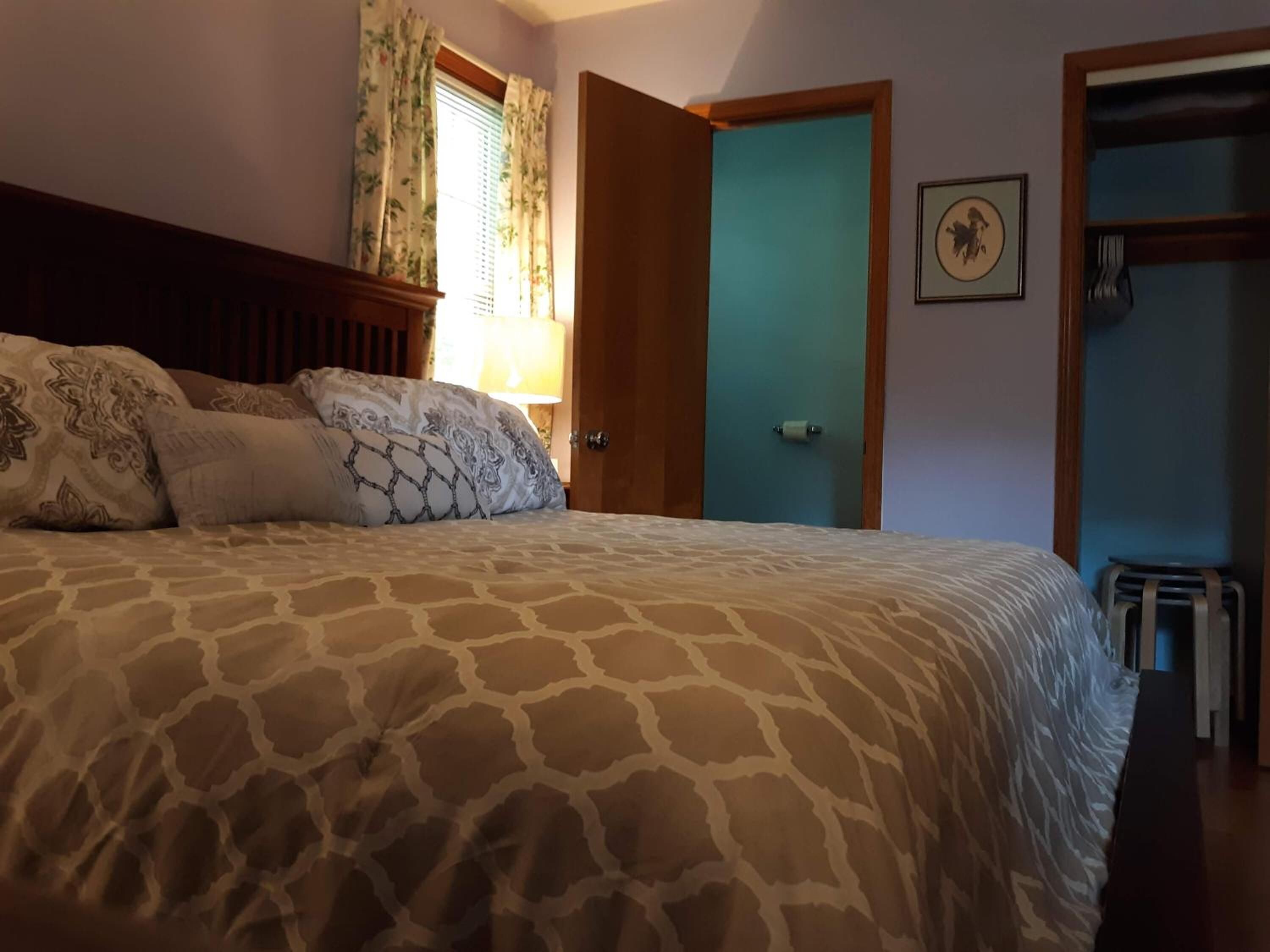 A bedroom with a patterned bedspread, a wooden headboard, a window with floral curtains, and a door leading to another room.