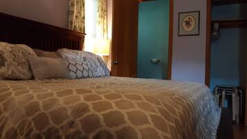 A bedroom with a patterned bedspread, a wooden headboard, a window with floral curtains, and a door leading to another room.