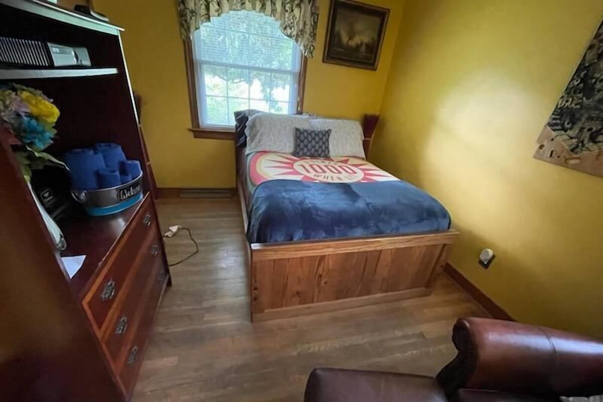 Une chambre à coucher avec un lit en bois, une commode, une fenêtre avec des rideaux et un tableau accroché au mur.