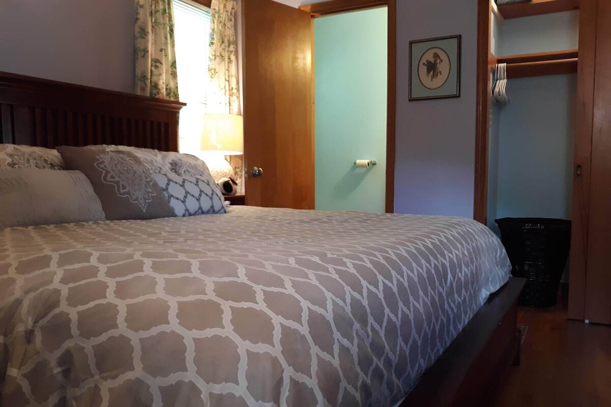 Une chambre avec un grand lit, une tête de lit en bois, une table de chevet avec une lampe, une armoire avec un miroir et un tableau encadré au mur.