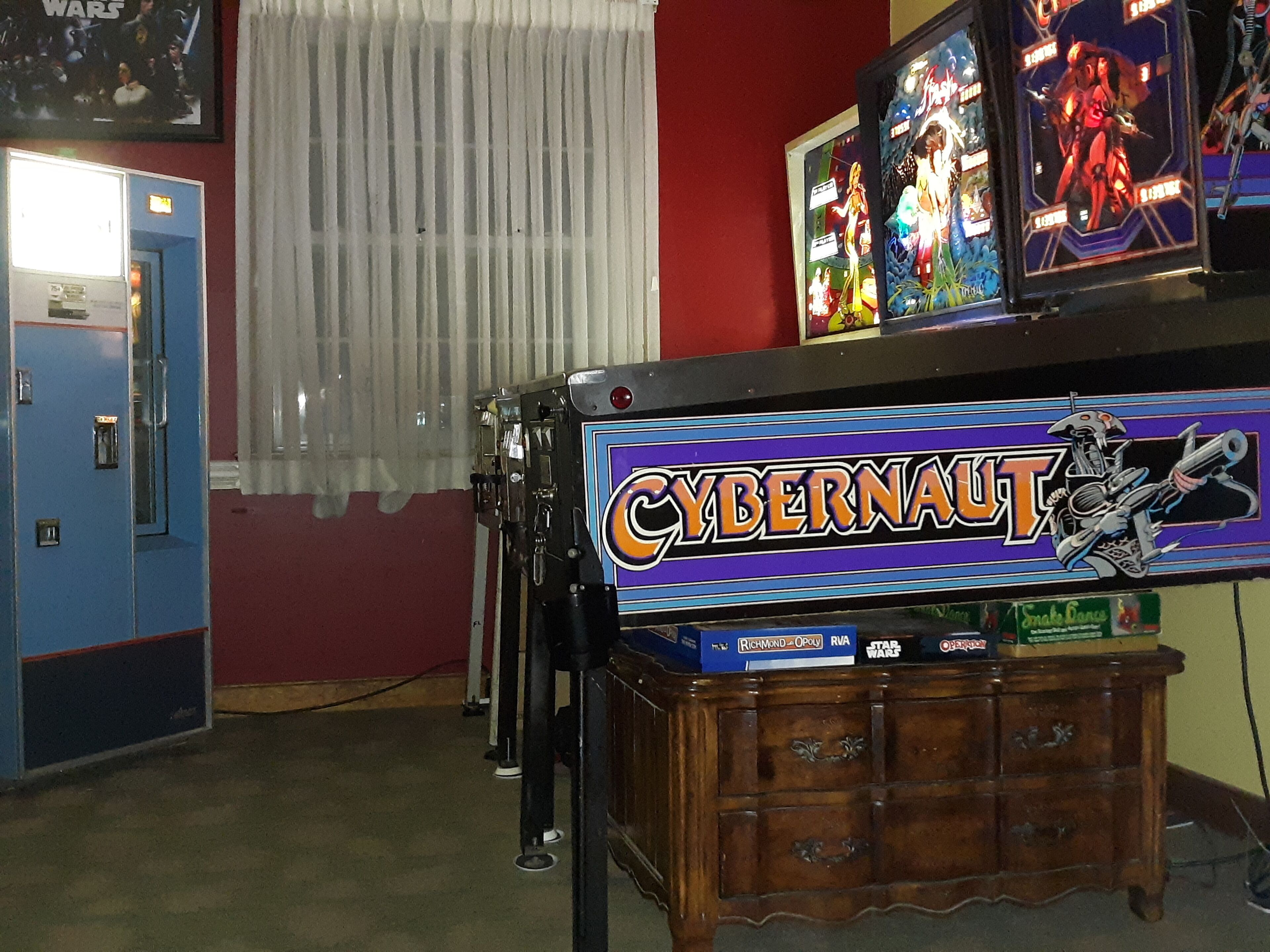 Ein Zimmer mit einer blauen Tür, einem Fenster mit weißen Vorhängen und einem Holfachschrank mit einem 'Cybernaut' Arcade-Spiel.