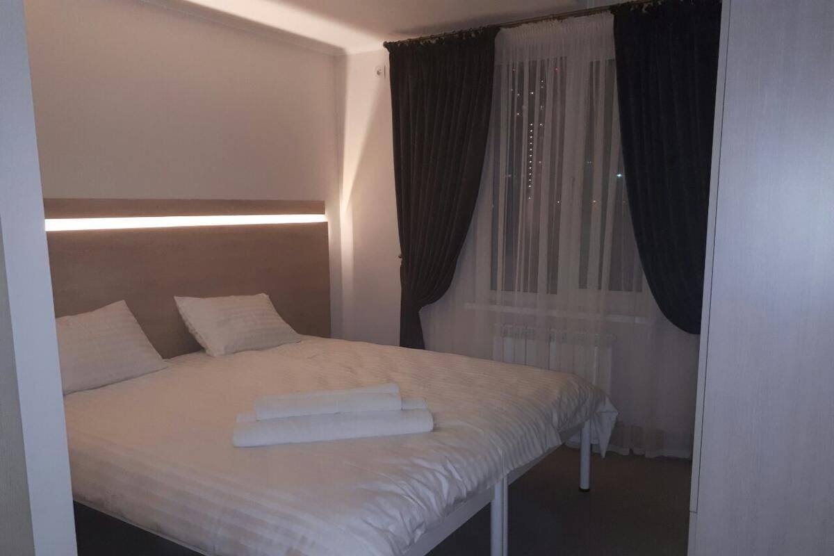 A neatly made bed with white linens and a folded towel, a bedside table, and a window with sheer curtains.