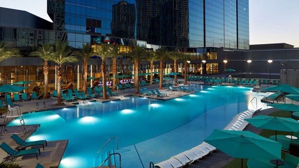 A large indoor swimming pool with lounge chairs and umbrellas, surrounded by palm trees and modern buildings.