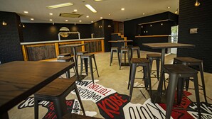 A modern restaurant interior with wooden tables and stools, a bar area, and a black wall with a glass display. - The Phat Packers - Hostel (Otari)