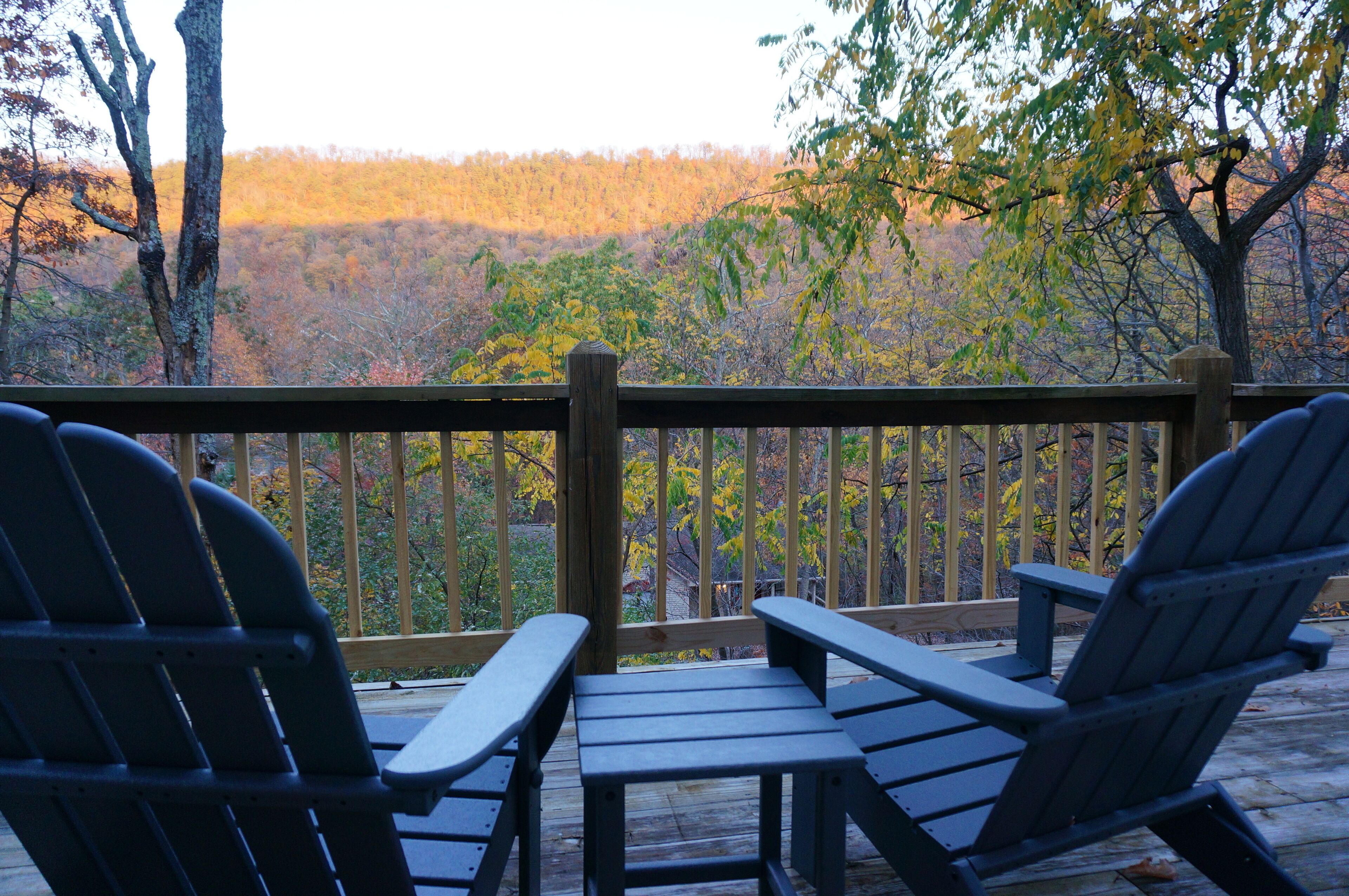 Um deck de madeira com duas cadeiras Adirondack e uma mesa para piquenique, com vista para uma área arborizada.