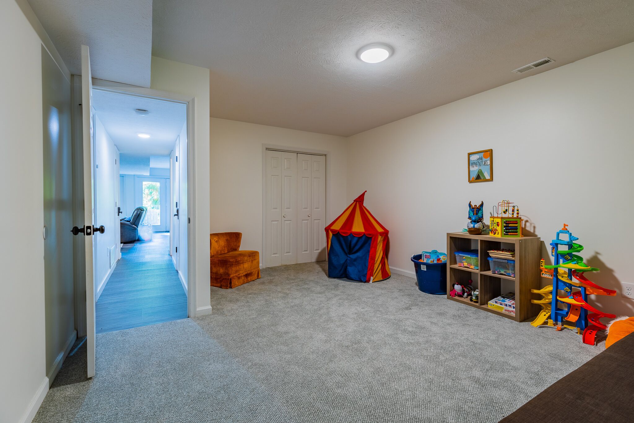 Une salle de jeux pour enfants avec un sol recouvert de moquette, une tente de jeu, une bibliothèque en bois et une porte donnant sur une autre pièce.
