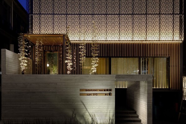 Un edificio moderno con fachada de madera, un letrero que dice 'SAGAKUKAN Hotel' y 'Sushi Syo', y un diseño de pared con motivos.