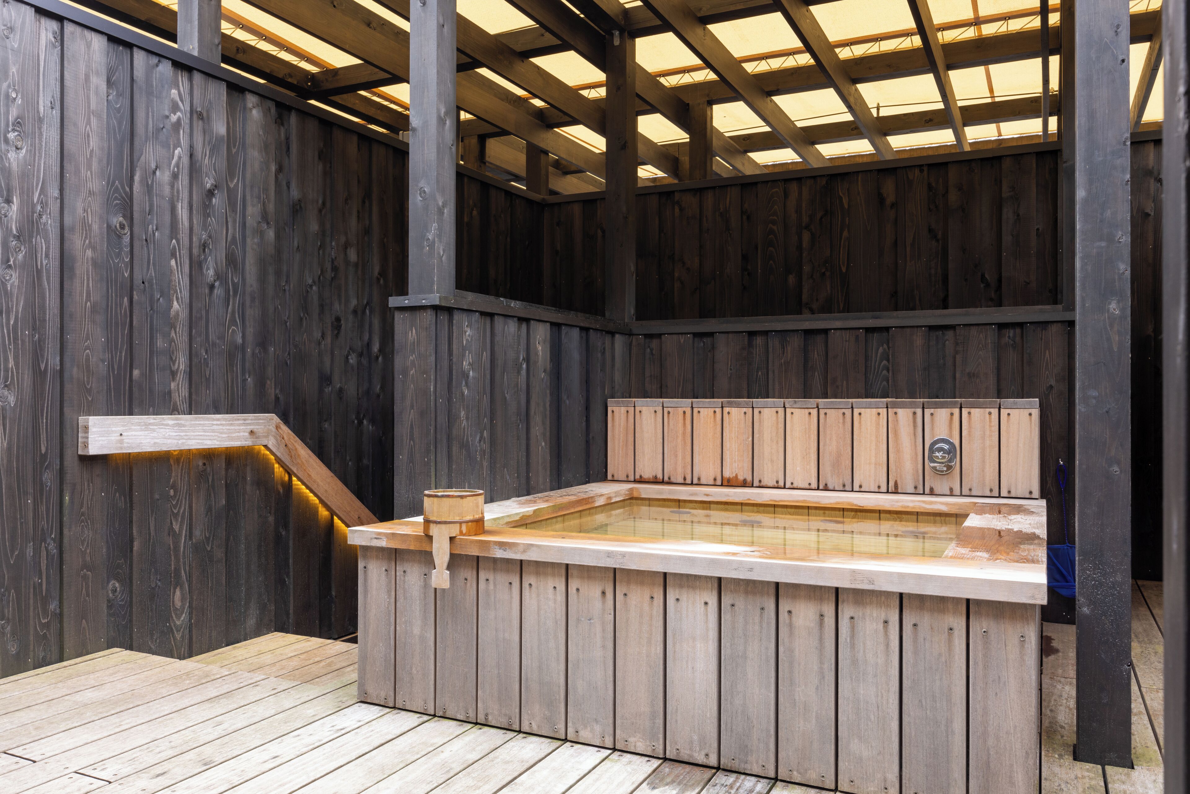 Un espacio tipo spa de madera con un jacuzzi, un muelle de madera y techo cubierto.