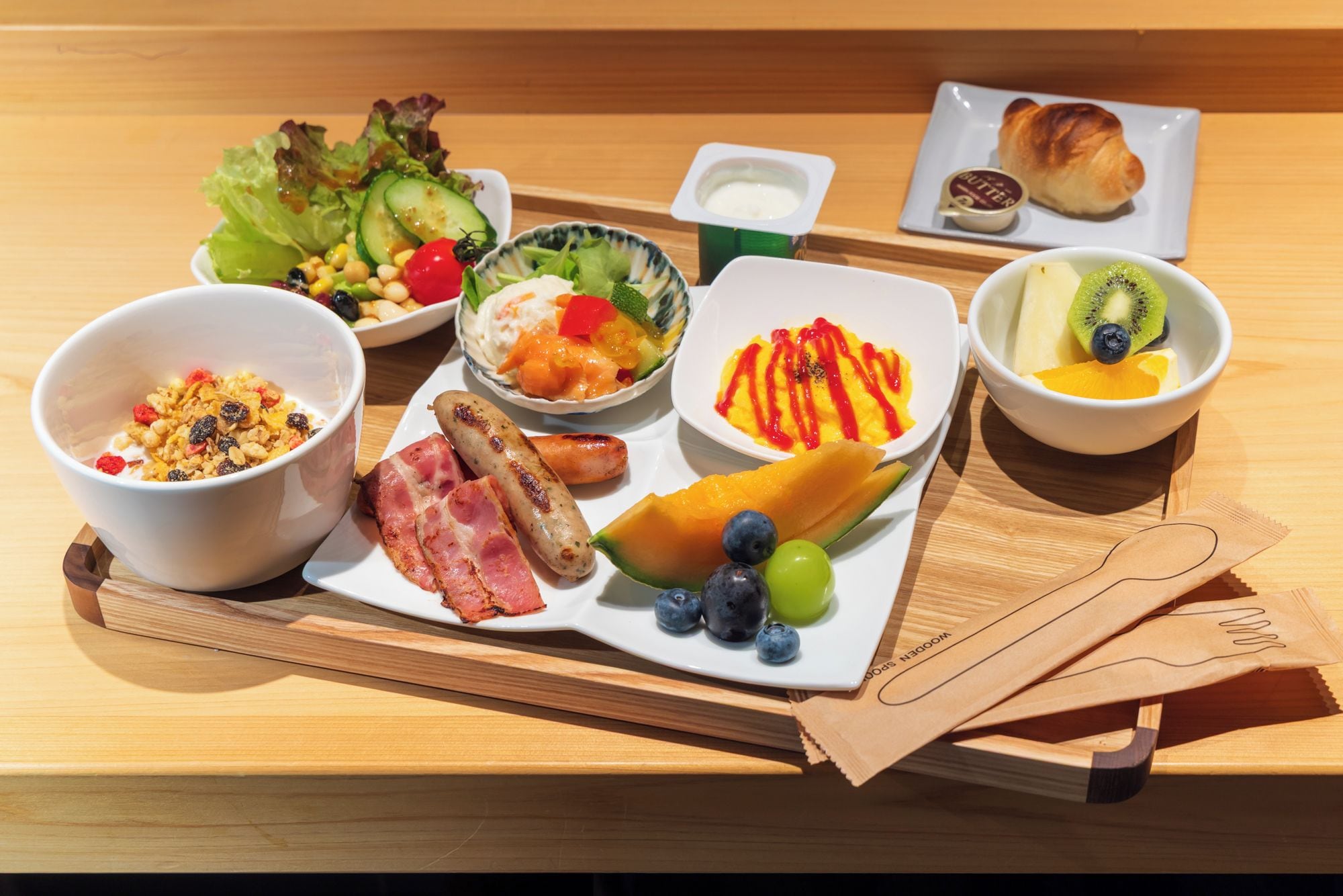 Bandeja de desayuno con diversos alimentos como cereal, salchicha, fruta y un croissant.
