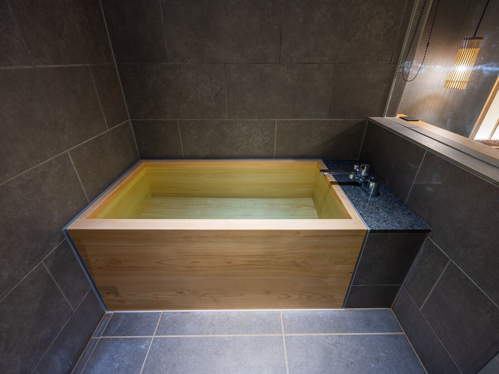 A modern bathroom with a wooden bathtub, dark tiles, and a small window.