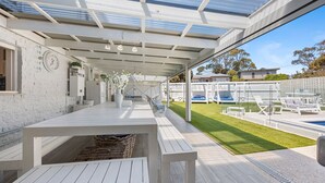 Ett takförsett utomhusområde med en pool, vita möbler och utsikt över trädgården.