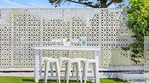 A white outdoor seating area with a table and chairs, set against a white lattice fence. - Santorini Beach House (Safety Beach)