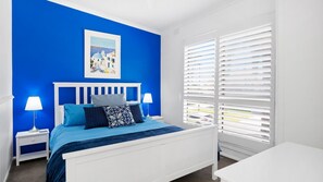 A bedroom with a blue wall, white bed, and a window with white blinds. - Santorini Beach House (Safety Beach)