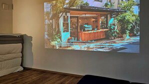A projection of a cafe with a wooden counter and outdoor seating, surrounded by trees and buildings.