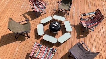 A wooden deck with a circular arrangement of white and striped lounge chairs, a central fire pit, and a small table.