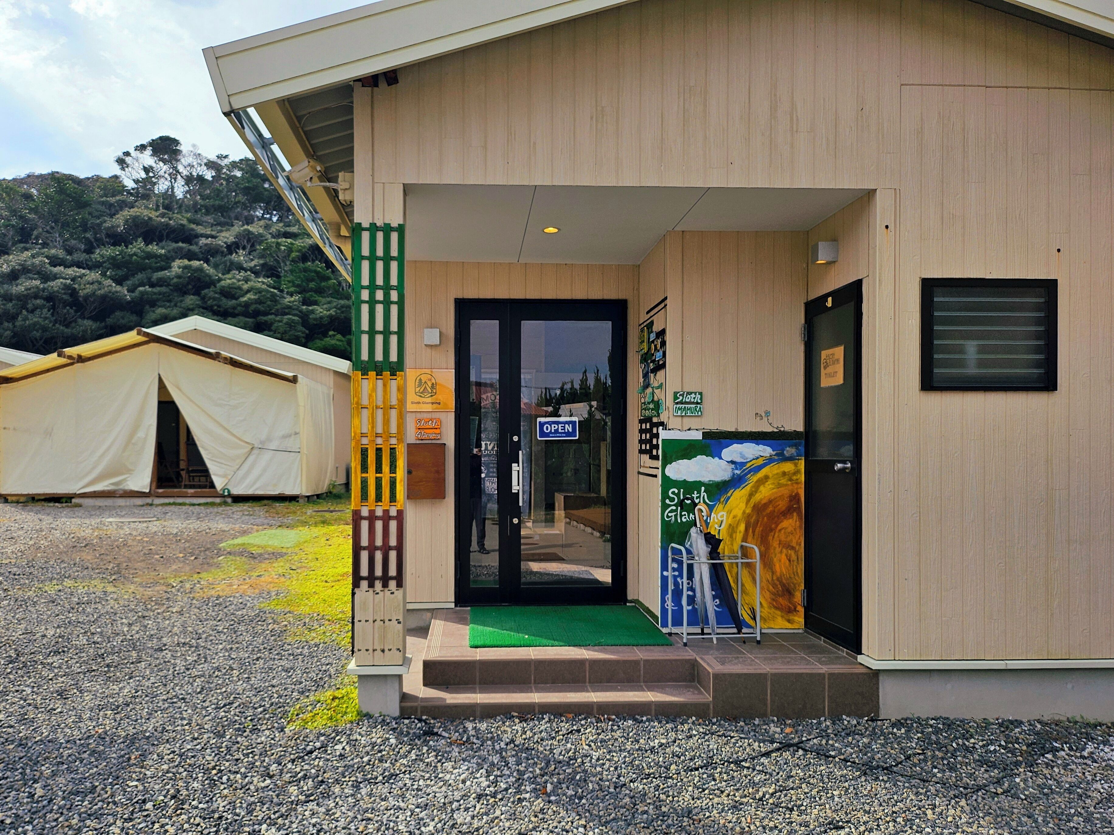 Un edificio moderno con fachada de madera, un felpudo verde en la entrada y un letrero que indica el nombre 'Sloth Glamping'.