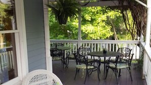 Varanda coberta com móveis de vime branco, incluindo cadeiras e uma mesa, e um corrimão de madeira.