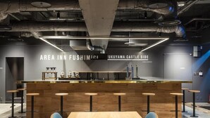 A modern bar area with a wooden bar counter and stools, a large table in the foreground, and a wall with signage reading "AREA INN FUSHIMI" and "UKUYAMA CASTLE SIDE CAFE LOUNGE". - Area Inn Fushimicho Fukuyama Castle Side 2-8 - Hostel (Fukuyama)