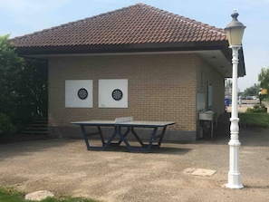 Un bâtiment avec un toit recouvert de tuiles, deux cibles de fléchettes accrochées au mur et une table placée devant.