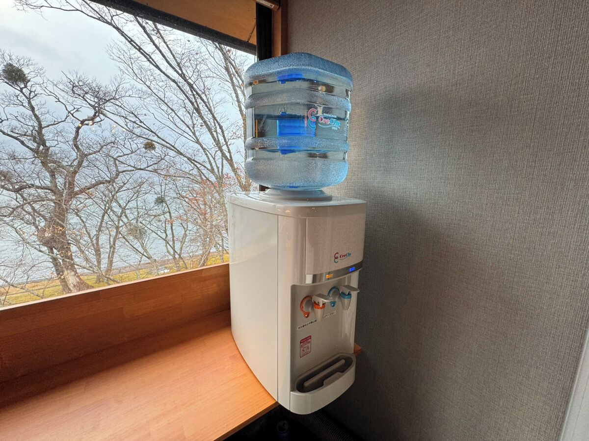 Dispensador de agua con un bidón grande encima, situado cerca de una ventana con vistas a árboles.