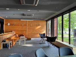 Un interior moderno con paneles de madera, una zona de estar con sofás y sillones, y ventanas grandes que ofrecen vistas a la vegetación exterior.