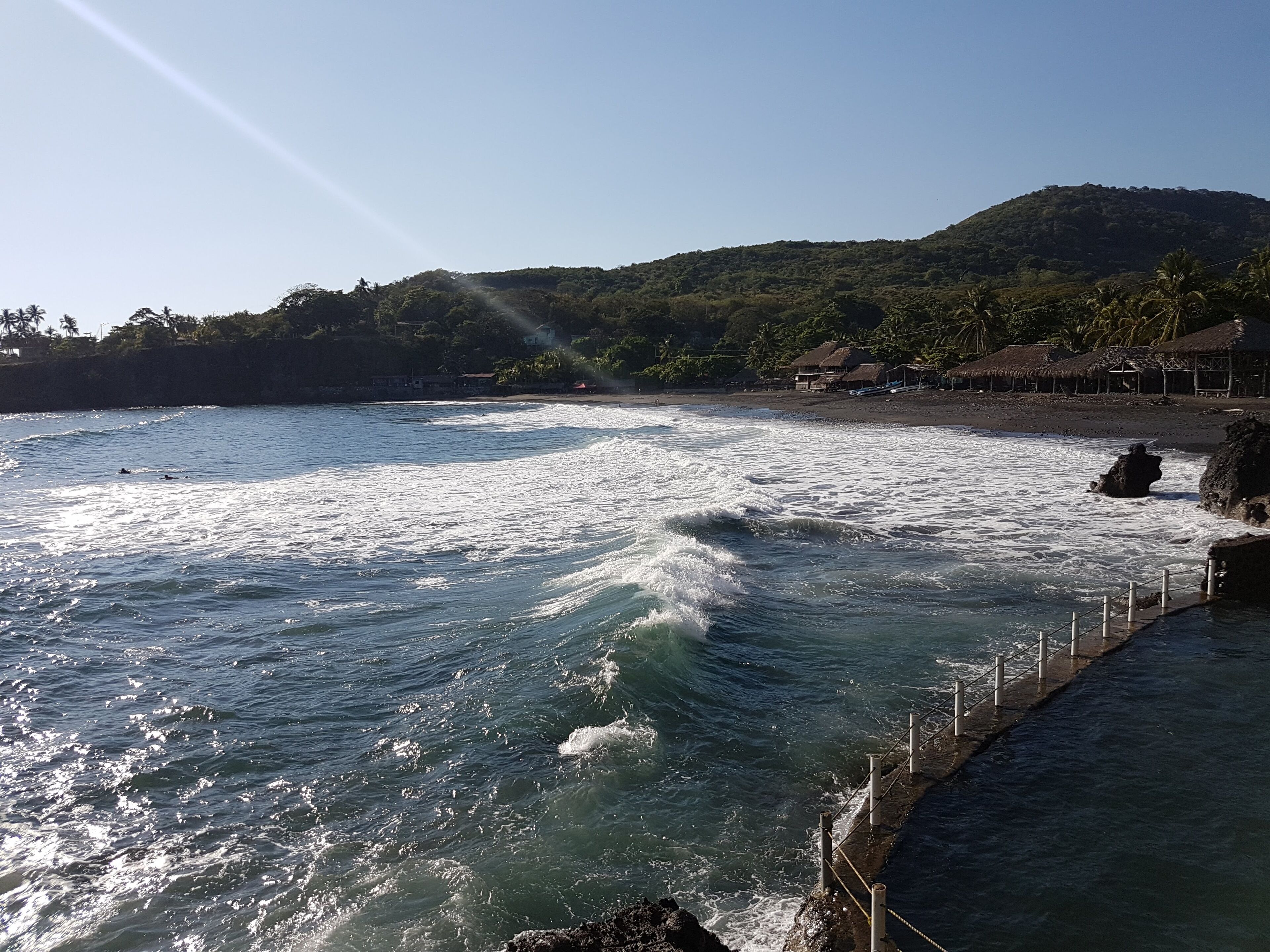 Paisaje costero con olas rompiendo sobre una costa rocosa, cielo despejado y un terreno montañoso al fondo.