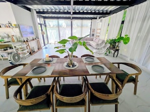 Un'area pranzo con un tavolo in legno apparecchiato per sei persone, circondato da sedie in vimini, un'ampia pianta in vaso e una cucina moderna sullo sfondo.