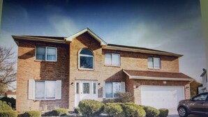 A two-story house with a garage, a front yard with bushes, and a car parked in the driveway. - Winding Meadows Home Away from Home (Bolingbrook)