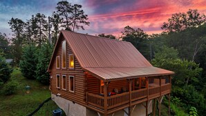 A wooden cabin with a balcony, surrounded by trees and a sunset sky.
