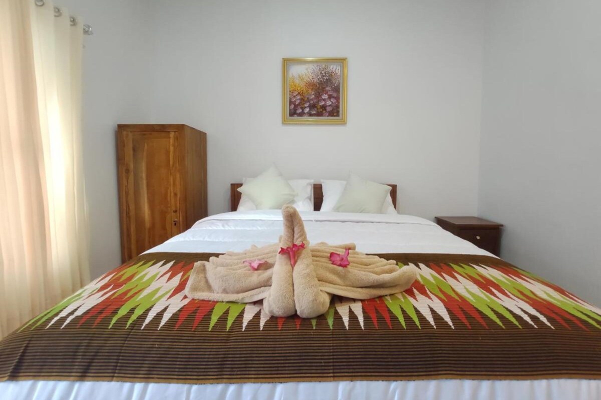 A neatly made bed with a patterned bedspread, a wooden wardrobe, and a painting on the wall.