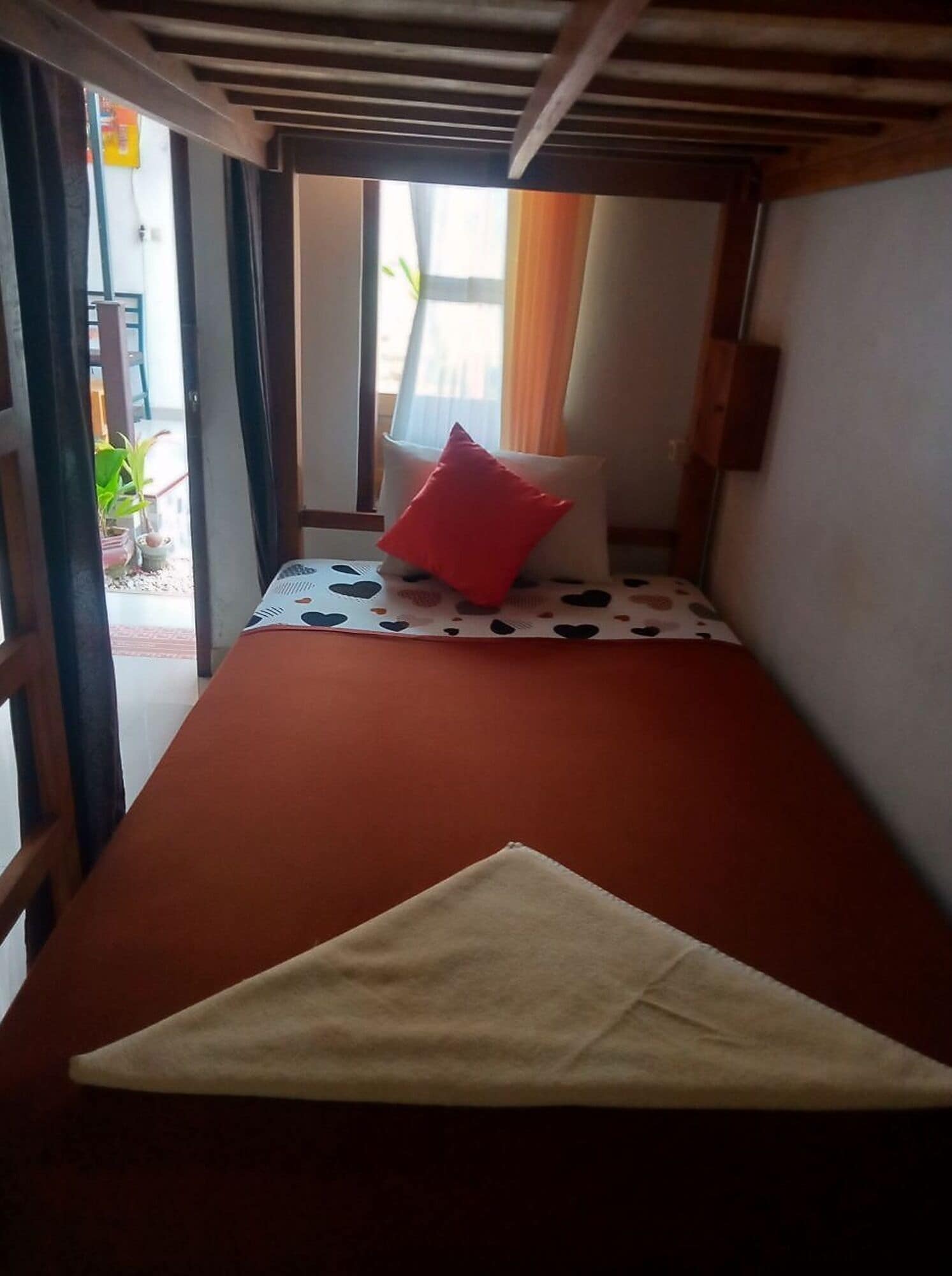 Uma cama de solteiro com colchão vermelho e travesseiro branco, uma mesa pequena com toalha de mesa estampada com corações e uma janela com cortinas.