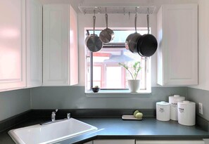 Una cocina con un lavabo blanco, encimera negra, ollas colgantes, una ventana con una planta y tres recipientes blancos etiquetados con té, harina y azúcar.