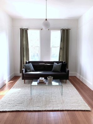 Una sala de estar con un sofá oscuro, una mesa de centro de vidrio y un tapete blanco. Hay dos ventanas con cortinas, y una lámpara colgante en el techo.