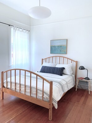 Un dormitorio con cama de madera, ropa de cama blanca, una mesita de noche con lámpara y un cuadro en la pared.