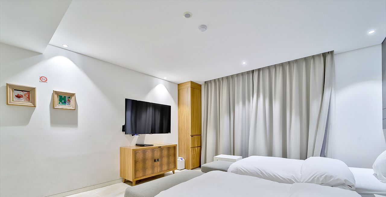 A modern hotel room with a large bed, a TV mounted on a wooden cabinet, a bedside table, and a large window with curtains.