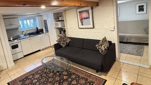 Ein kompakter Wohnbereich mit einer dunkelgrauen Couch, einem Glastisch und einem gemusterten Teppich. Im Hintergrund ist eine Küche mit weißen Schränken und einer Mikrowelle erkennbar.