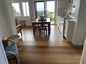 Un coin repas avec une table et des chaises en bois, un fauteuil en osier avec un coussin, et une vue sur l’extérieur à travers une porte vitrée.