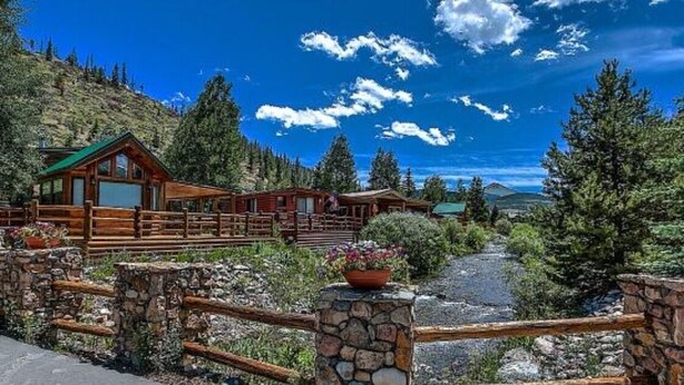 A wooden lodge with a stone entrance gate, surrounded by lush greenery and a clear blue sky. - Homey Hideaway Chalet in family friendly Tiger Run, mins from Breckenridge (Breckenridge)