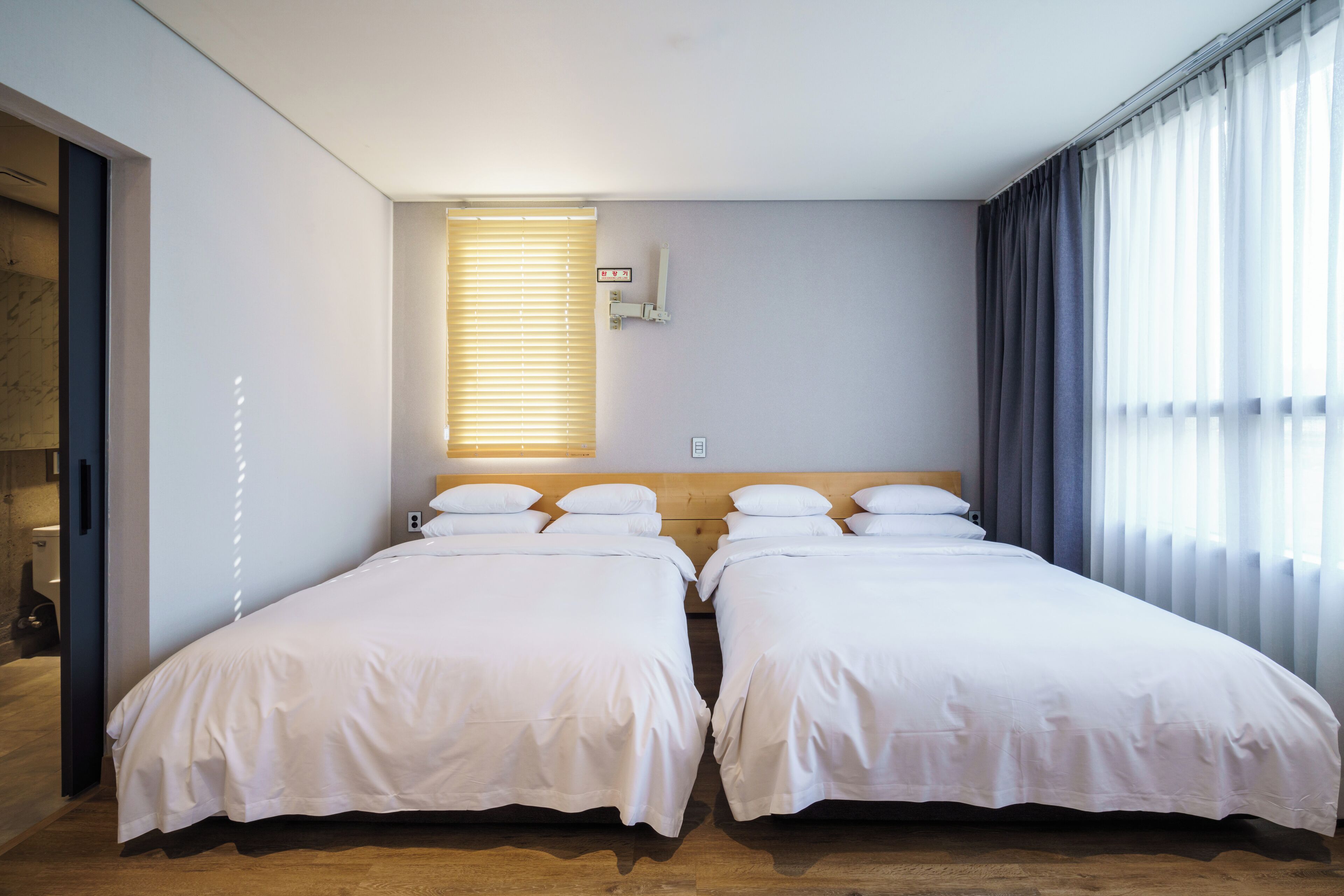 A hotel room with two beds, a wooden headboard, a window with blinds, and a wall-mounted light fixture.