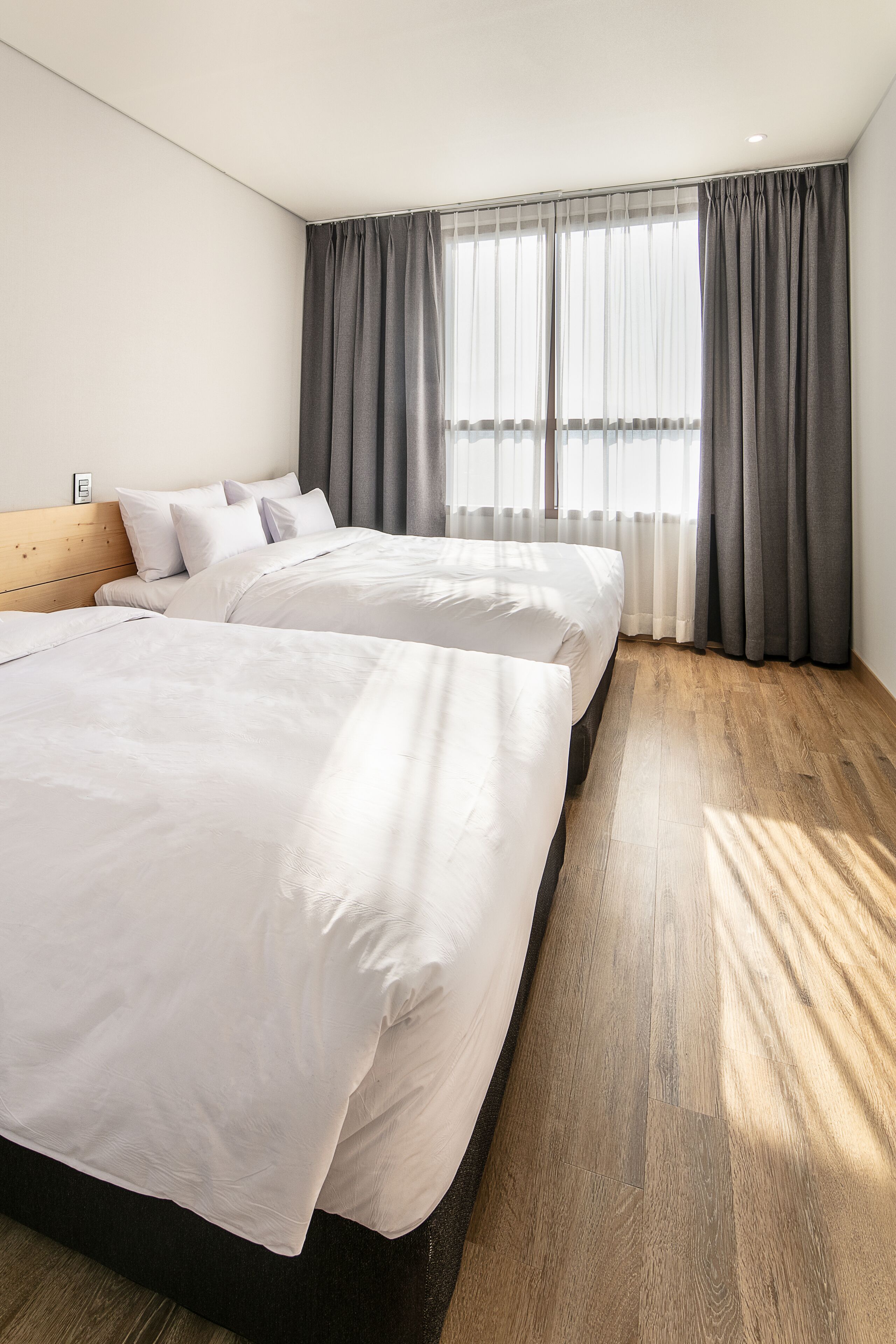 Una habitación de hotel moderna con dos camas, piso de madera y ventanas amplias con cortinas grises.