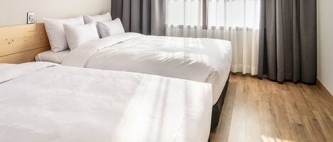 A modern hotel room with two beds, a wooden floor, and large windows with grey curtains.
