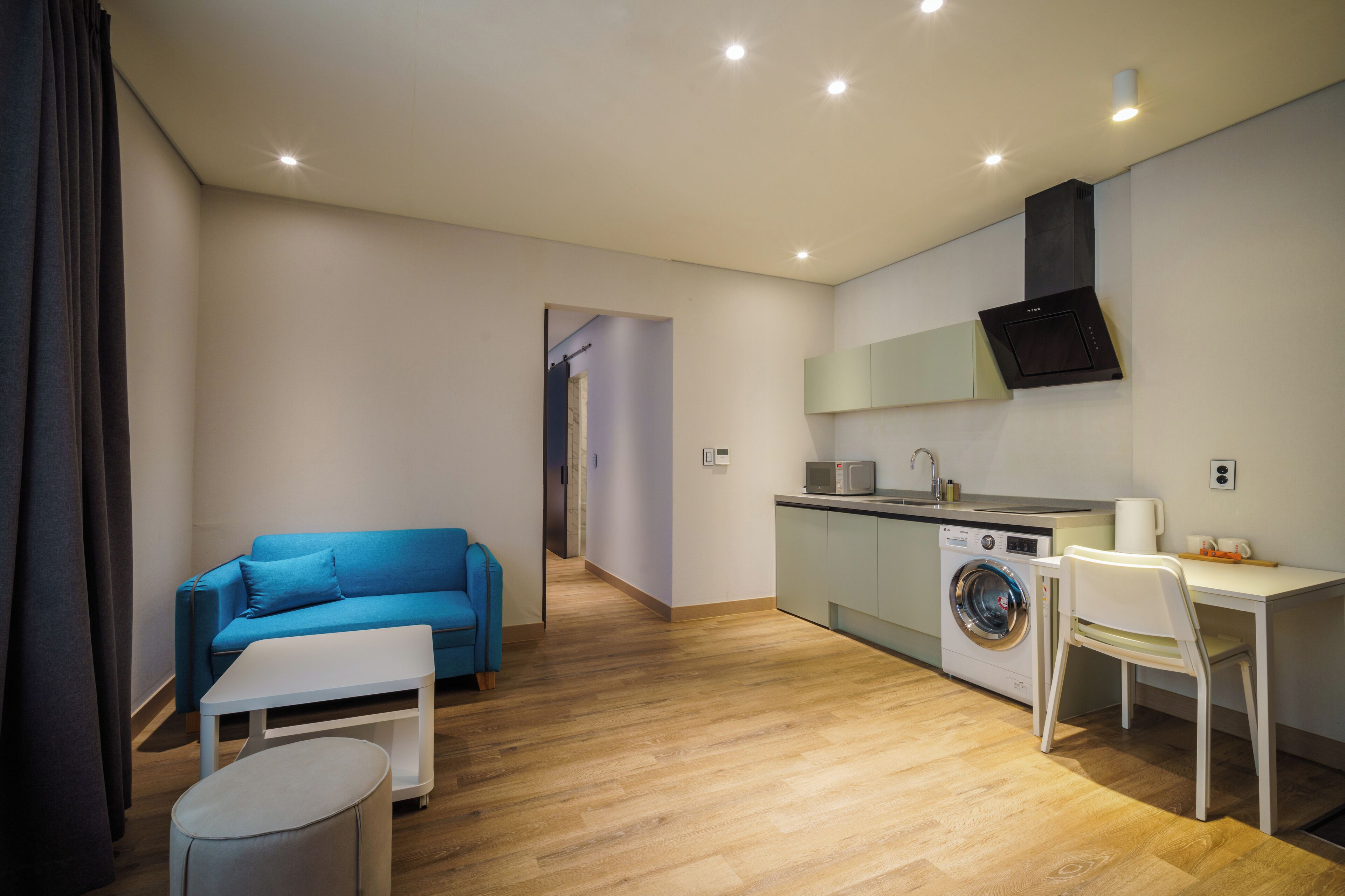 A modern apartment with a living area featuring a blue sofa, a small white table, and a round ottoman. A kitchen with a washing machine and a microwave is visible, along with a dining area with a white chair.