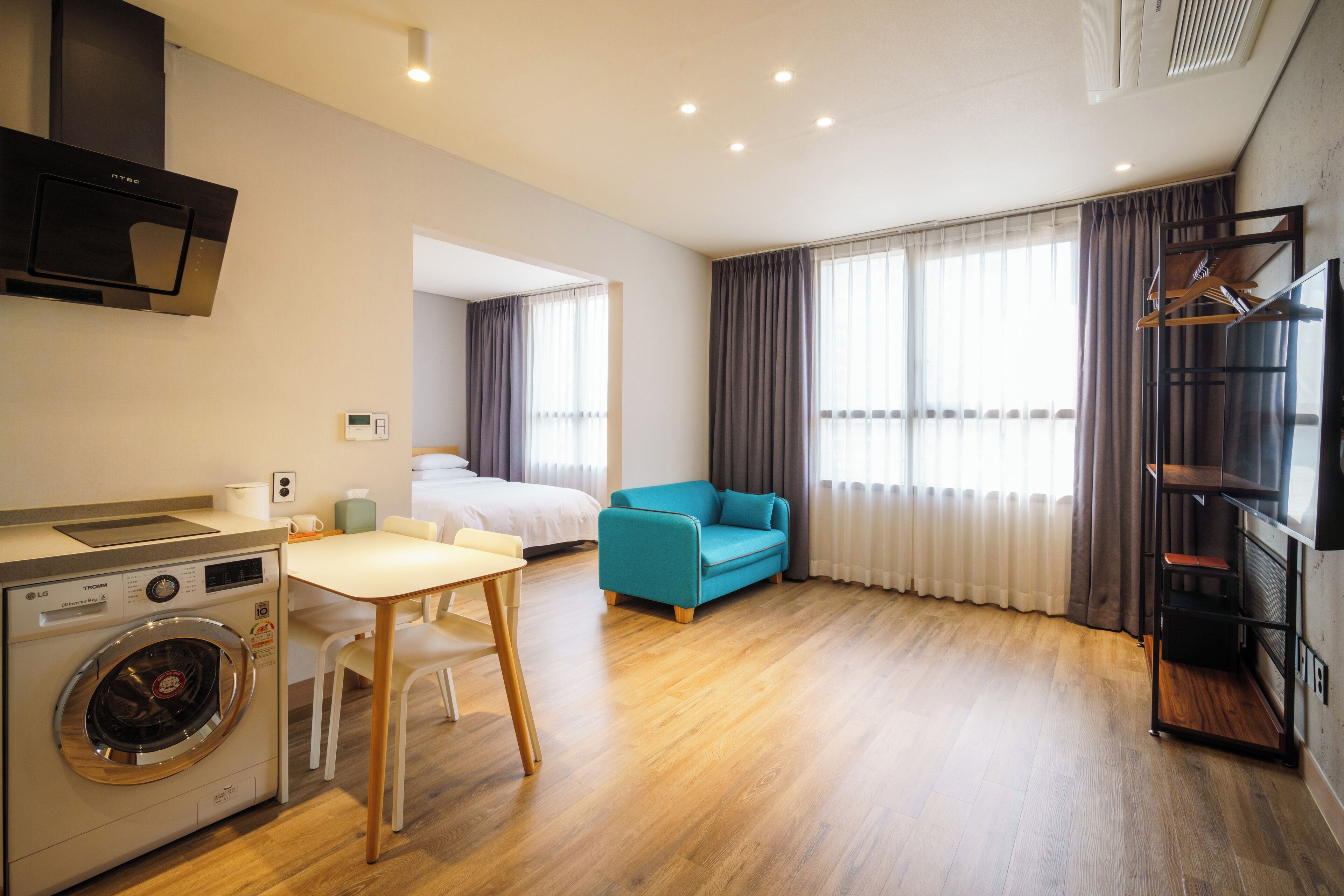 Una habitación de hotel moderna con lavadora, zona de comedor, sofá, televisor y ventanas grandes con cortinas.