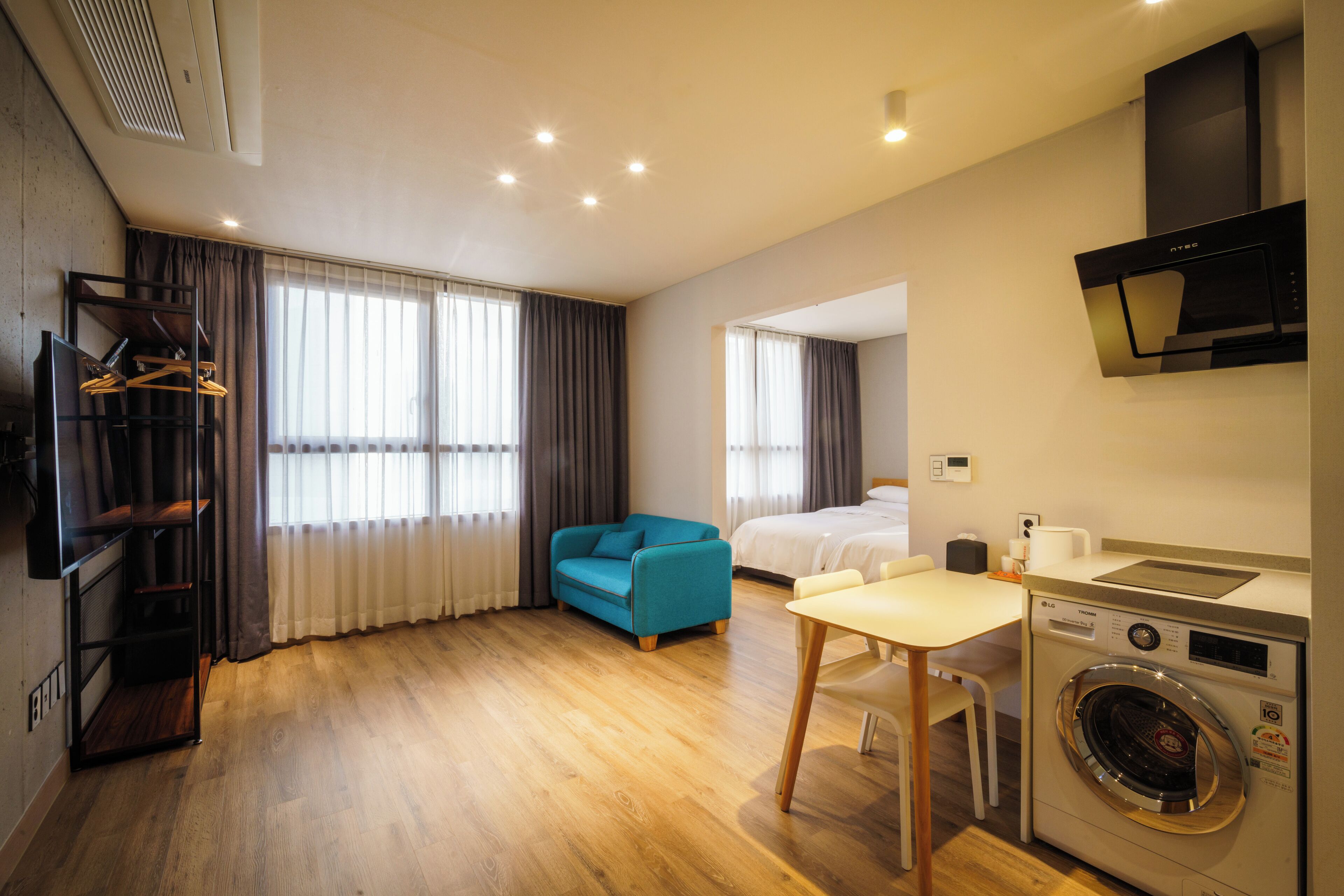 A modern hotel room with wooden flooring, a blue sofa, a bed, a washing machine, and a TV mounted on the wall.