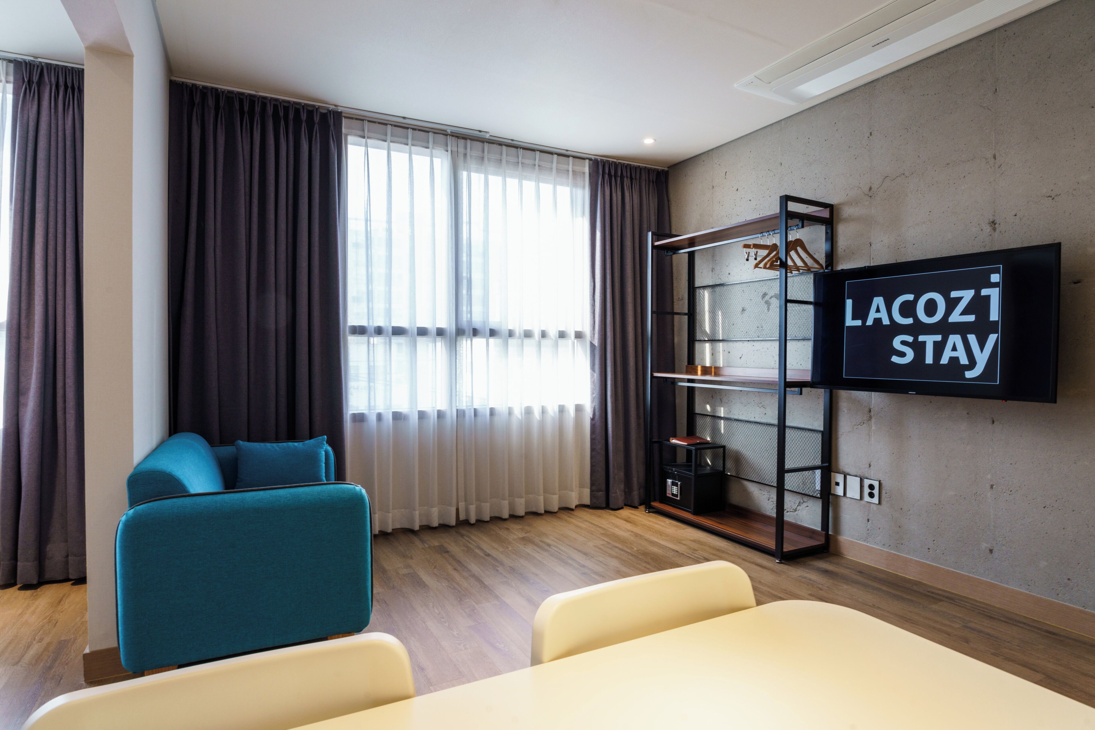 A modern hotel room with a sofa, a TV mounted on the wall, a wooden floor, and a large window with curtains.