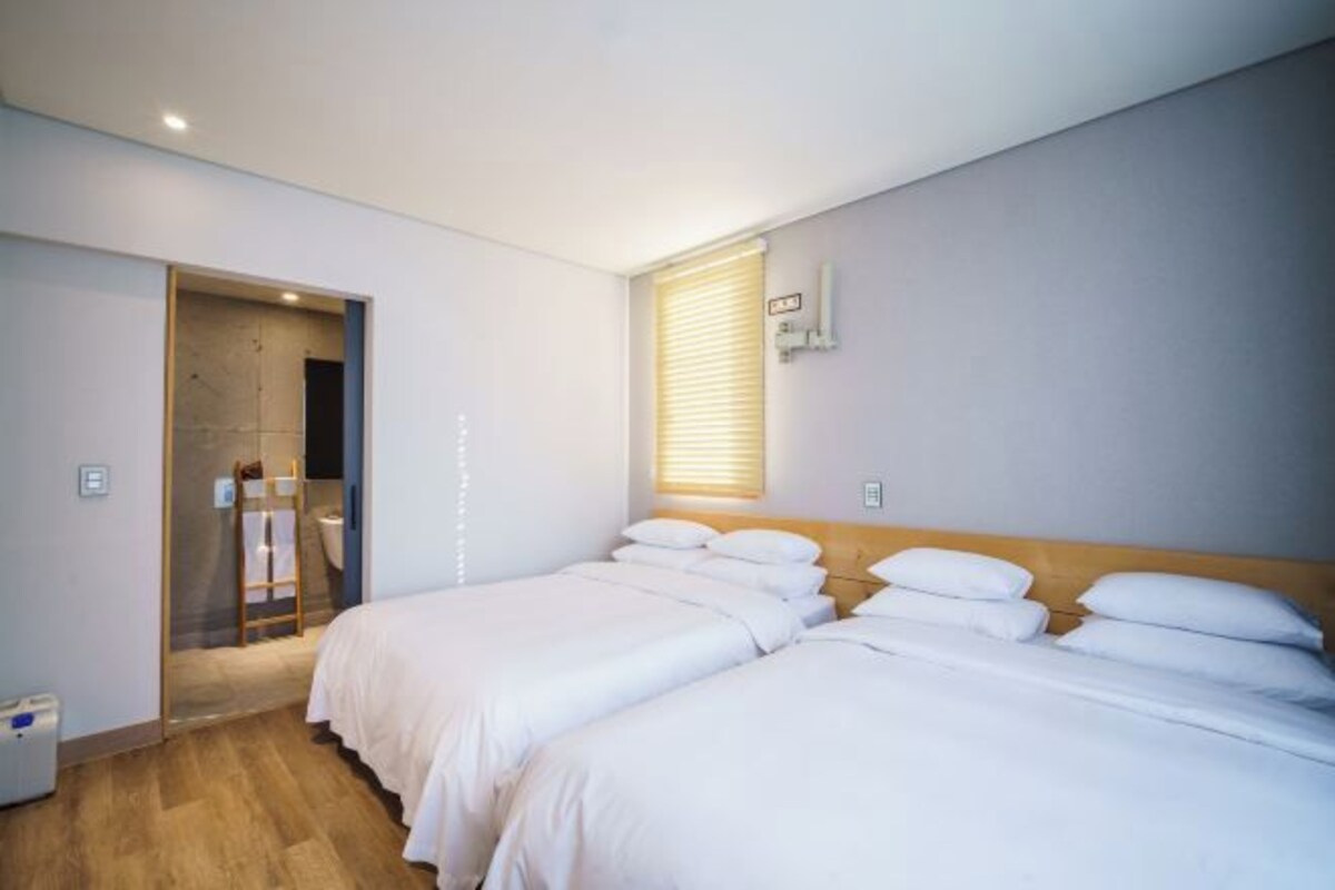 Habitación de hotel con dos camas, suelo de madera, baño con toallero y ventana con persianas.