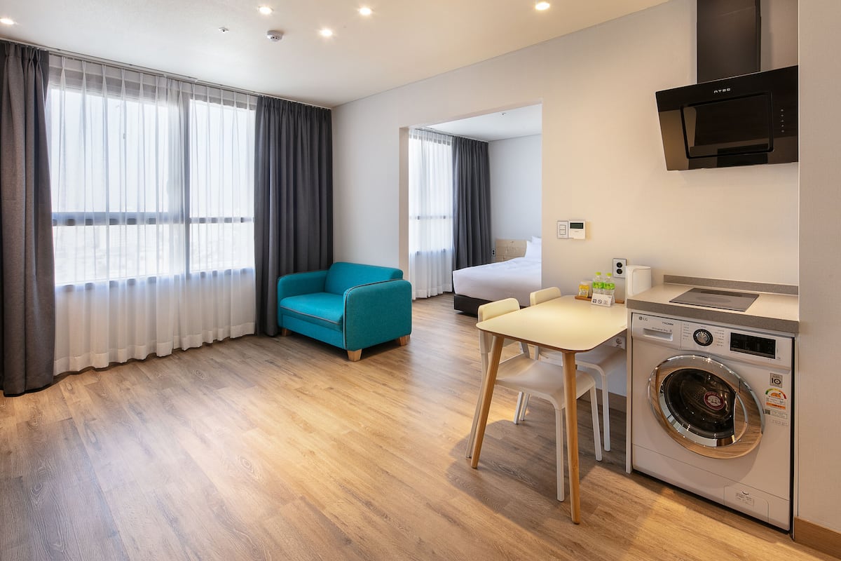 Una moderna camera d'albergo con pavimento in legno, un divano color petrolio, un piccolo tavolo da pranzo, una lavatrice e un letto con vista attraverso tende leggere.