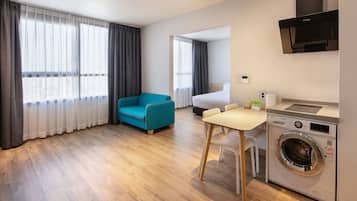 A modern hotel room with a wooden floor, a teal sofa, a small dining table, a washing machine, and a bed with a view through sheer curtains.