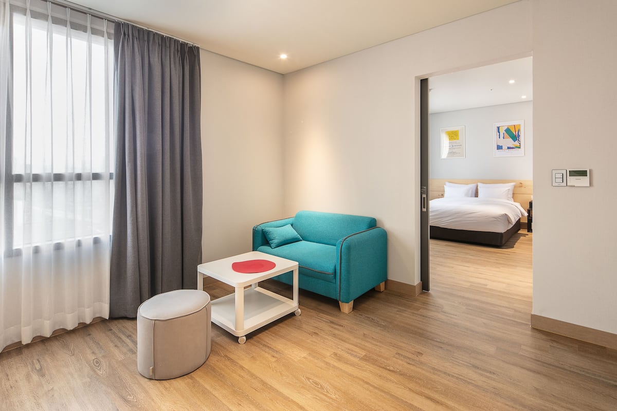 Una moderna camera d'hotel con un divano color acquamarina, un piccolo tavolo bianco con un oggetto rosso, un poggiapiedi rotondo e un pavimento in legno.