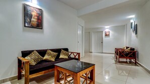 A living room with a sofa, a glass-top coffee table, and a patterned cushion. There is a door leading to another room and a small table with a chair in the background. - Itsy Hotels Dew Dreams (Aluva)